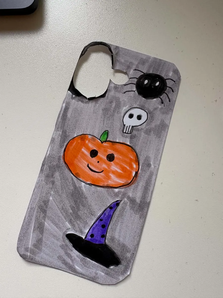 A hand-drawn Halloween phone case insert on grey marker paper with a pumpkin, spider, skull and witch hat, cut to phone shape with a camera hole